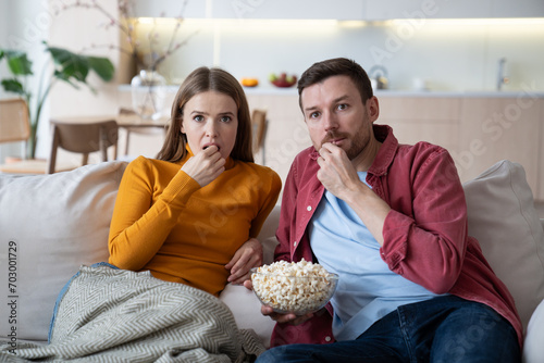 Intrigued couple engrossed in watching interesting fascinating film  video  program  show on TV  sitting on couch  eating popcorn. Spending lockdown  covid isolation  weekend  pastime together at home