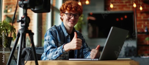Happy ginger teenager influencer recording blog, talking to camera on tripod, seated at table with laptop. Smiling teen displaying thumbs up.