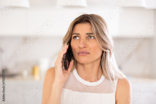 Irritated blonde talking on phone while sitting on the kitchen table