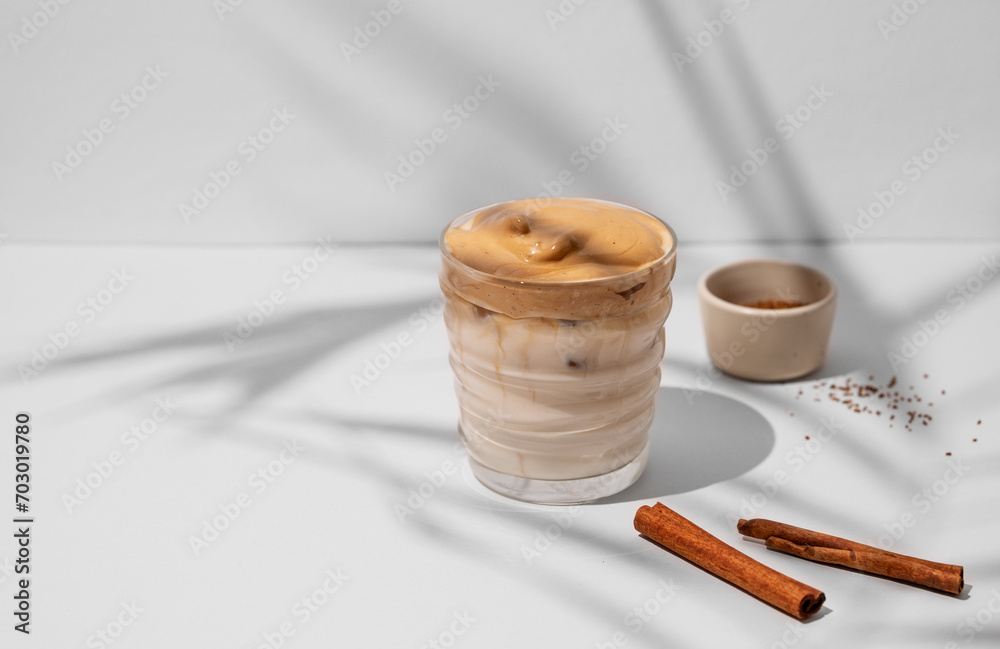 Dalgona coffee. Whipped instant iced coffee with milk in a glass with cinnamon sticks on gray background with shadow. Dalgona coffee. Whipped instant iced coffee with milk in a glass with cinnamon sticks on gray background with shadow.