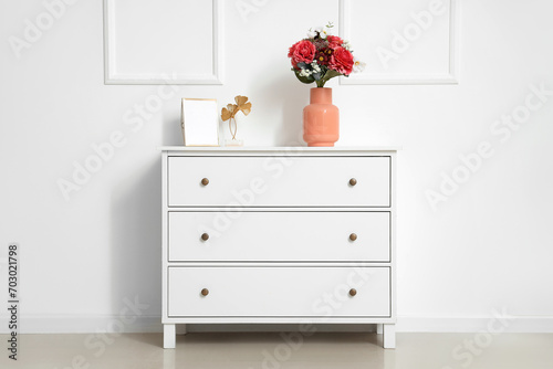 Vase with beautiful artificial flowers on chest of drawers near white wall