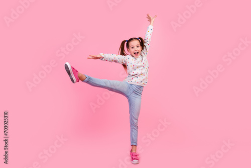 Full length photo of small girl wear stylish sweatshirt jeans raising leg up stand on tiptoe dancing isolated on pink color background