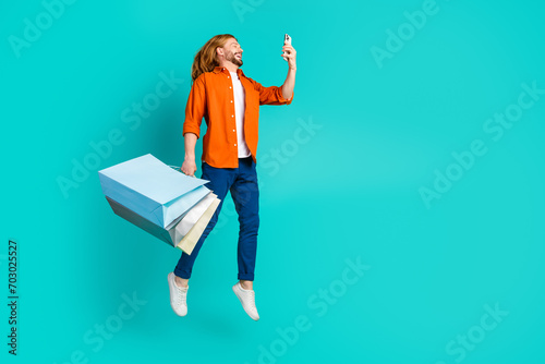 Full size photo of clever guy dressed blue jeans flying hold new clothes look at smartphone isolated on turquoise color background