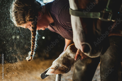 Quarter horse getting hoof shaped by a female farrier with two pigtail braids in a dusty stall in an old wooden barn.
