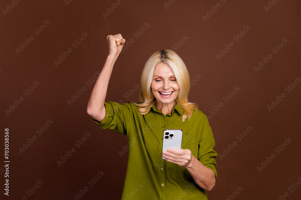 Photo of overjoyed woman with blond hair dressed khaki shirt look at smartphone win lottery isolated on dark brown color background Photo of overjoyed woman with blond hair dressed khaki shirt look at smartphone win lottery isolated on dark brown color background
