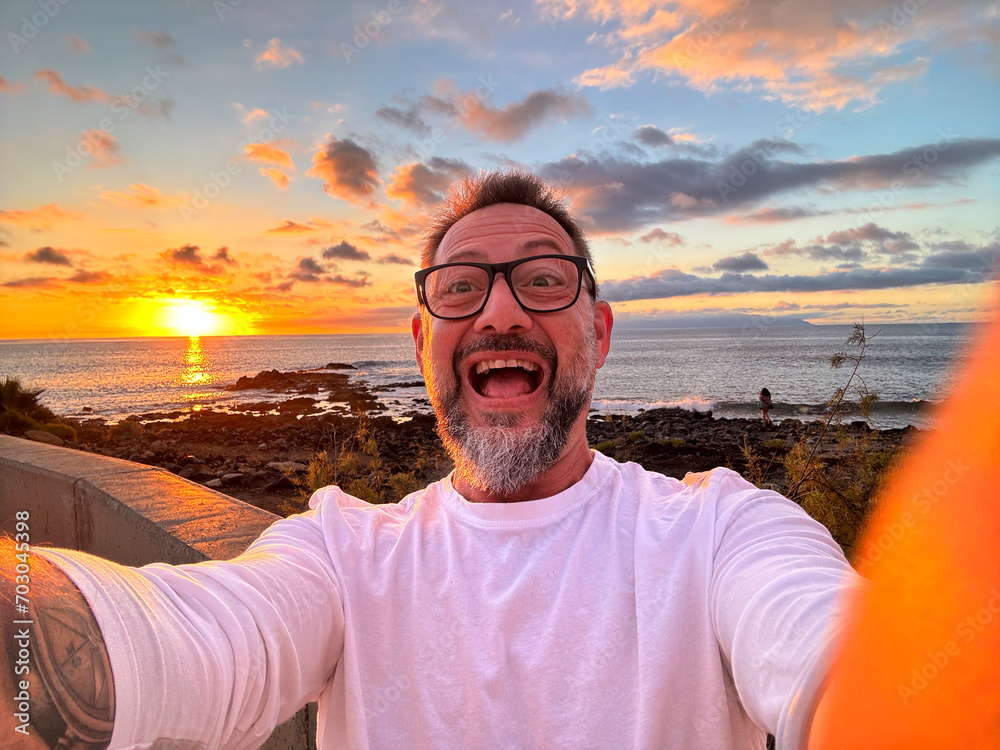Joyful happy expression portrait of man taking selfie picture during sunset time in tropical island travel wanderlust destination. People and outdoor leisure activity in summer holiday vacation. Phone