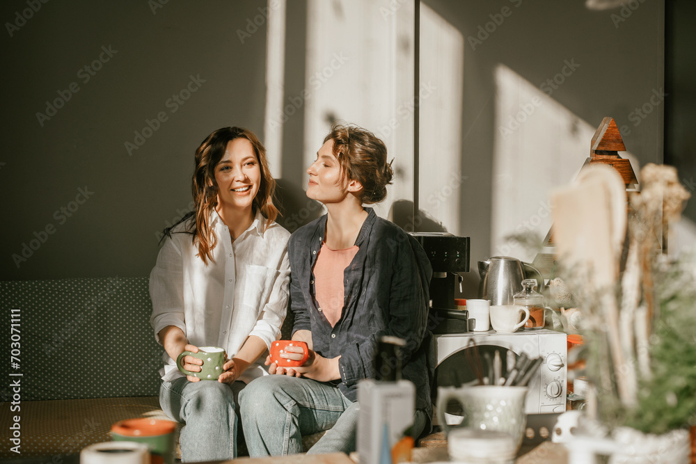 Cozy morning: girls friends talking over a cup of coffee in a bright kitchen Two women spend time peacefully on a sunny day with mugs of tea - obrazy, fototapety, plakaty 