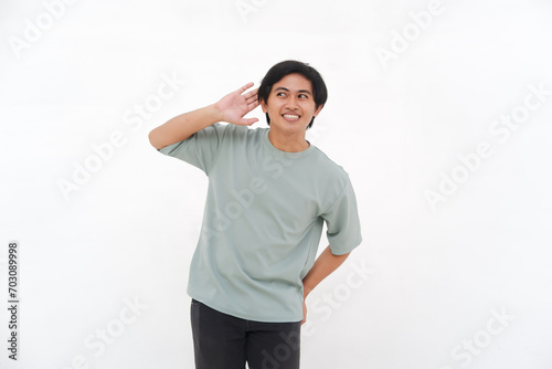 A young Asian man, an employee in a tshirt, is seen enthusiastically listening with his hand to his ear against a white background.