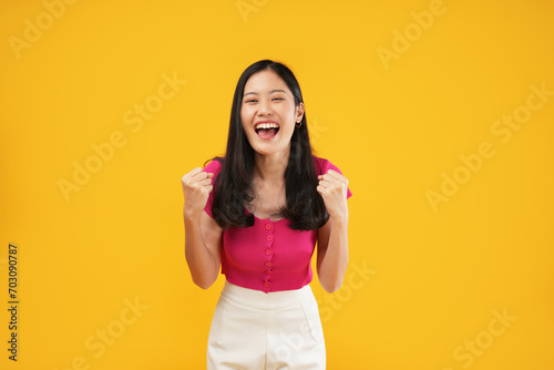 Portrait of a young Asian woman casually dressed woman screaming yes and clenching her fists, winner's gesture concept. Isolated on a yellow background. 