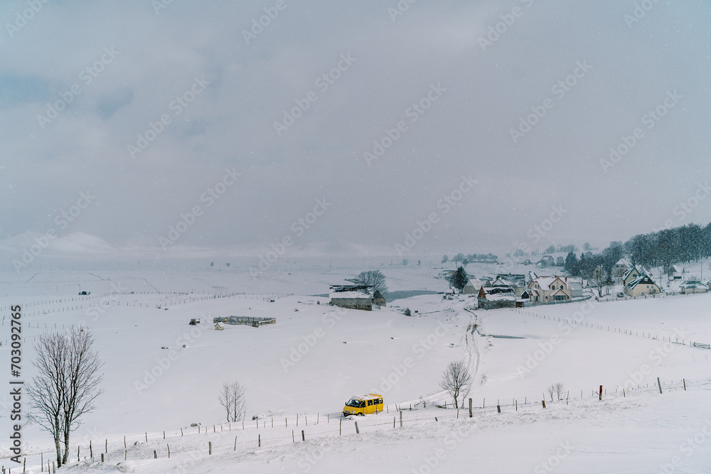 Yellow minibus drives along a snowy road in a small village