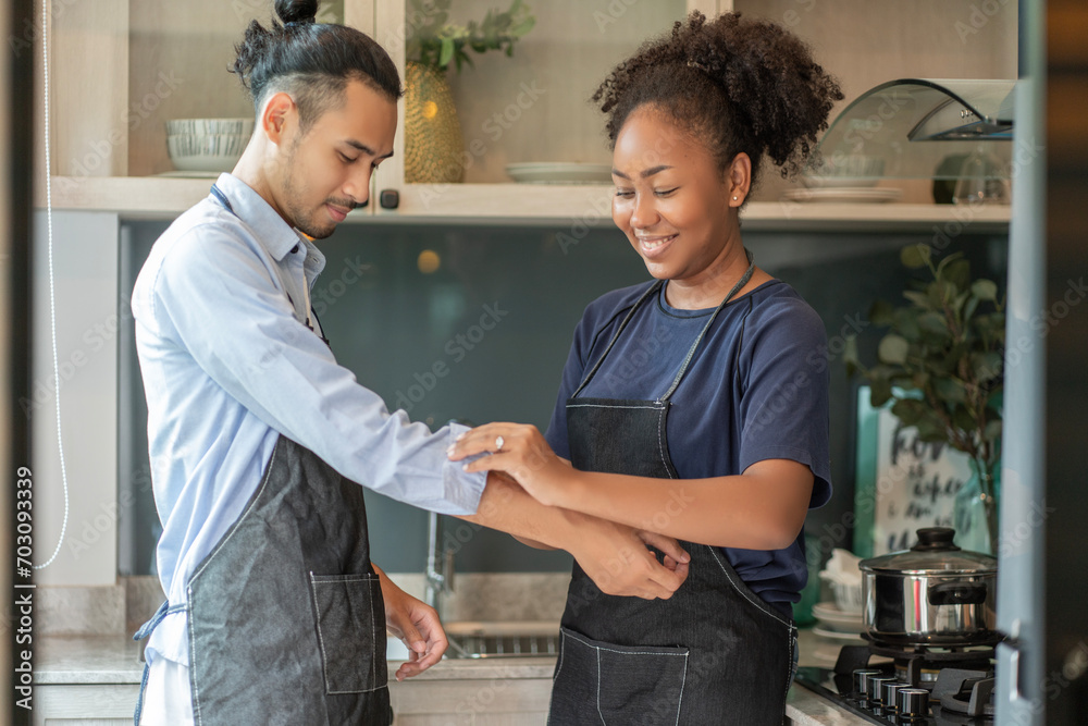 Fototapeta premium Black African woman, wife taking care of Asian husband, getting her husband dressed and rolling up her sleeves in the kitchen at home.