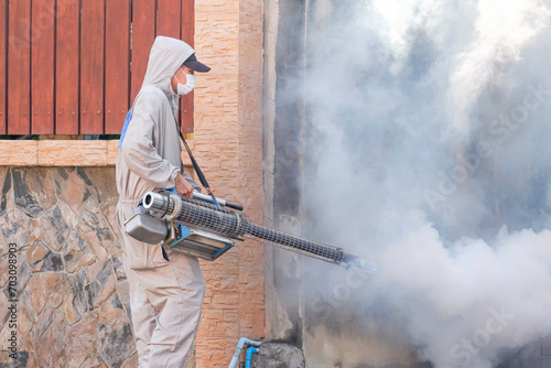 Outdoor healthcare worker using fogging machine spraying chemical to removal mosquitoes and control dengue fever epidemic in abandoned area next to the fence wall of house photo