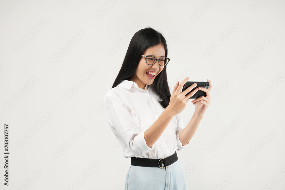 This portrait of a young Asian woman wearing a white shirt and glasses, with a positive expression playing video games on a modern smartphone, isolated on a white background. Modern technology concept This portrait of a young Asian woman wearing a white shirt and glasses, with a positive expression playing video games on a modern smartphone, isolated on a white background. Modern technology concept