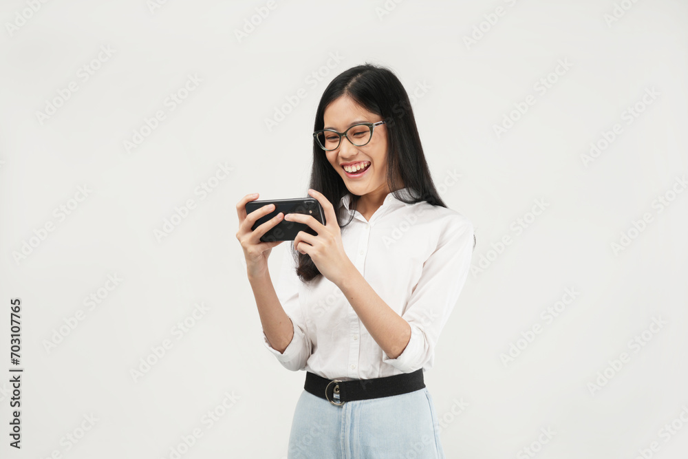 This portrait of a young Asian woman wearing a white shirt and glasses, with a positive expression playing video games on a modern smartphone, isolated on a white background. Modern technology concept This portrait of a young Asian woman wearing a white shirt and glasses, with a positive expression playing video games on a modern smartphone, isolated on a white background. Modern technology concept