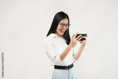 This portrait of a young Asian woman wearing a white shirt and glasses, with a positive expression playing video games on a modern smartphone, isolated on a white background. Modern technology concept