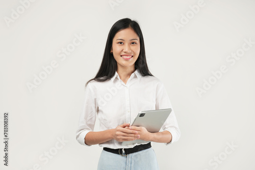 Photo of an Asian woman 20s standing and holding a tablet, dressed a white shirt and isolated on a white background. 
