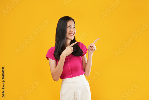 Portrait of a young Asian woman with satisfied expression, points away with both fore fingers for copy space, isolated on a yellow background.