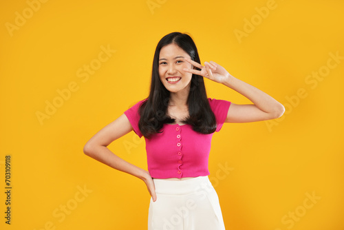 Portrait of a young Asian lady showing a v-sign advertisement  Wearing a pink t-shirt. Isolated on a yellow background. Peace concept. 