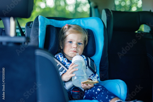 Adorable baby girl with blue eyes sitting in car safety seat. Toddler child going on family vacations and jorney. Smiling happy child during traffic jam, drinking milk from bottle and eating bisquit photo