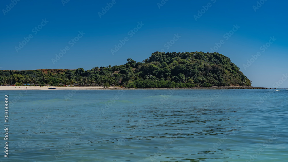 The beautiful tropical island  with lush green vegetation. Tiny silhouettes of people, moored boats, and houses of local residents are visible on the sandy beach. Clear blue sky, turquoise ocean. 