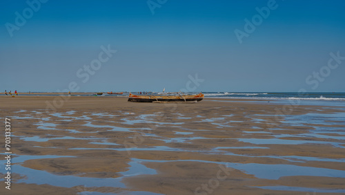 Low tide in the ocean. Traditional Malagasy wooden pirogue boats are located on the exposed seabed. Tiny silhouettes of people, a sailboat in the distance.The blue sky is reflected in puddles of water