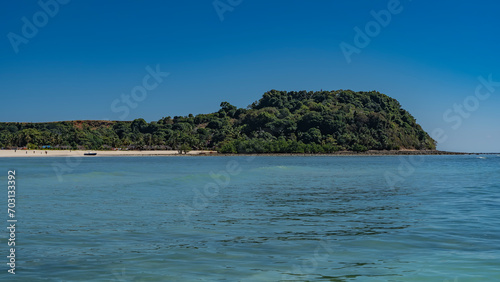 The beautiful tropical island with lush green vegetation. Tiny silhouettes of people, moored boats, and houses of local residents are visible on the sandy beach. Clear blue sky, turquoise ocean. 