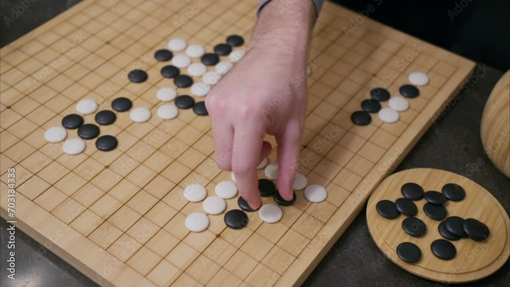 45 degree overhead angle of hand playing white stone in Go board game with tiles already on grid and black pieces in corner 45 degree overhead angle of hand playing white stone in Go board game with tiles already on grid and black pieces in corner