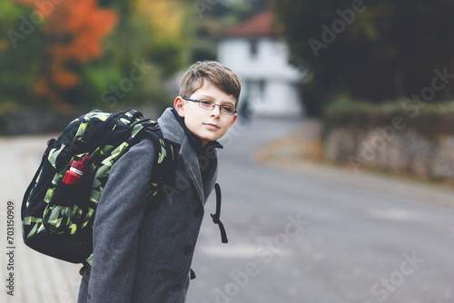 Happy kid boy with glasses and backpack or satchel. Schoolkid in stylish fashon coan on the way to middle or high school on cold autumn day. Healthy child outdoors on the street, on rainy day. photo