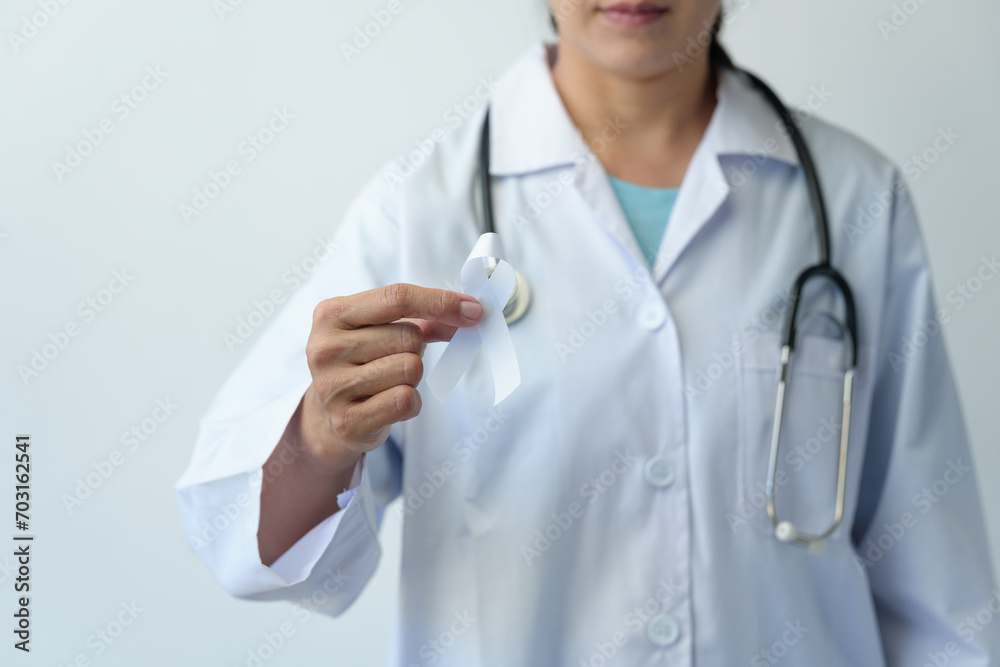 Image of a woman's hand in a doctor's uniform and stethoscope holding a white ribbon representing the struggle for life. Treated from lung cancer World cancer day concept, health insurance, prevention Image of a woman's hand in a doctor's uniform and stethoscope holding a white ribbon representing the struggle for life. Treated from lung cancer World cancer day concept, health insurance, prevention