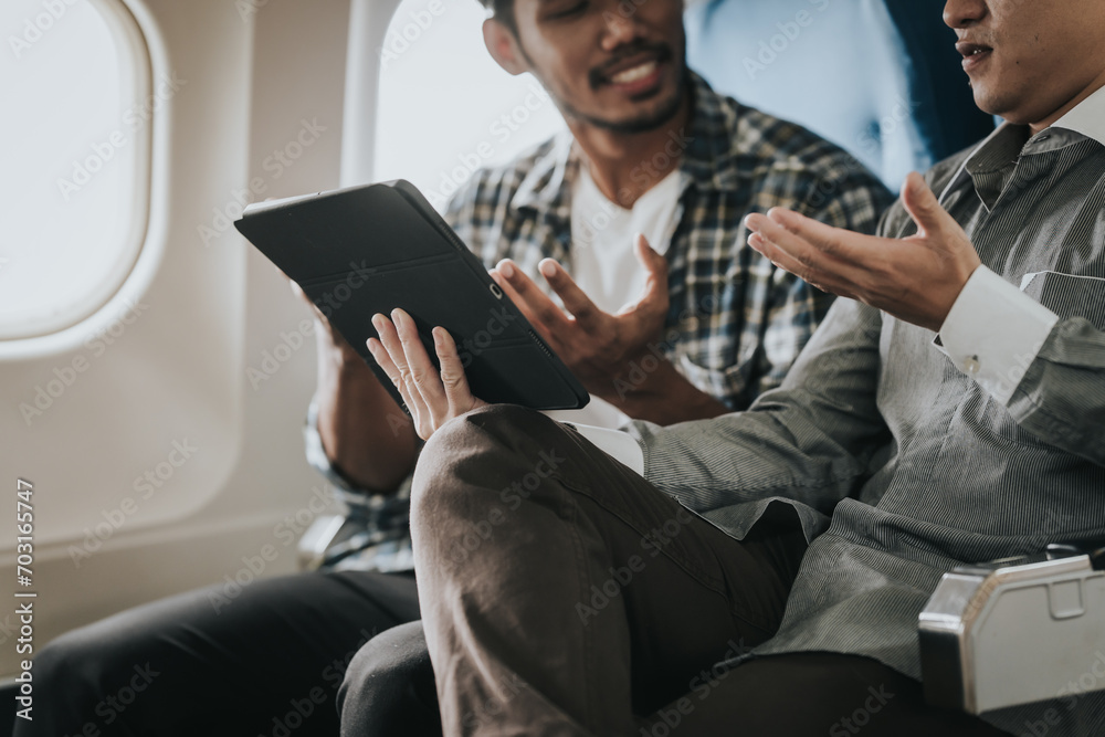 two Asian men on an airplane, looking at a tablet together, possibly friends choosing a location or planning their travel itinerary, sharing a cheerful moment.