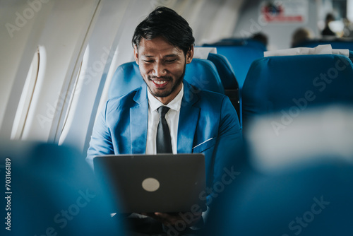 Asian businessman working on his laptop while seated in an airplane, smiling, possibly engaged in work or leisure during his flight.