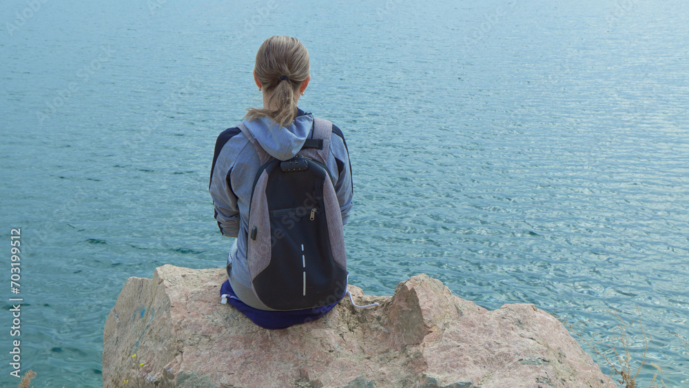 Fototapeta premium A woman tourist with a backpack on her shoulders sits on the top of a mountain and looks at the ocean.