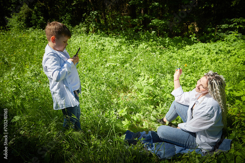 A young teenage son takes pictures of his mother on a cell phone in nature. Mom and boy having fun in the grass in a clearing photo