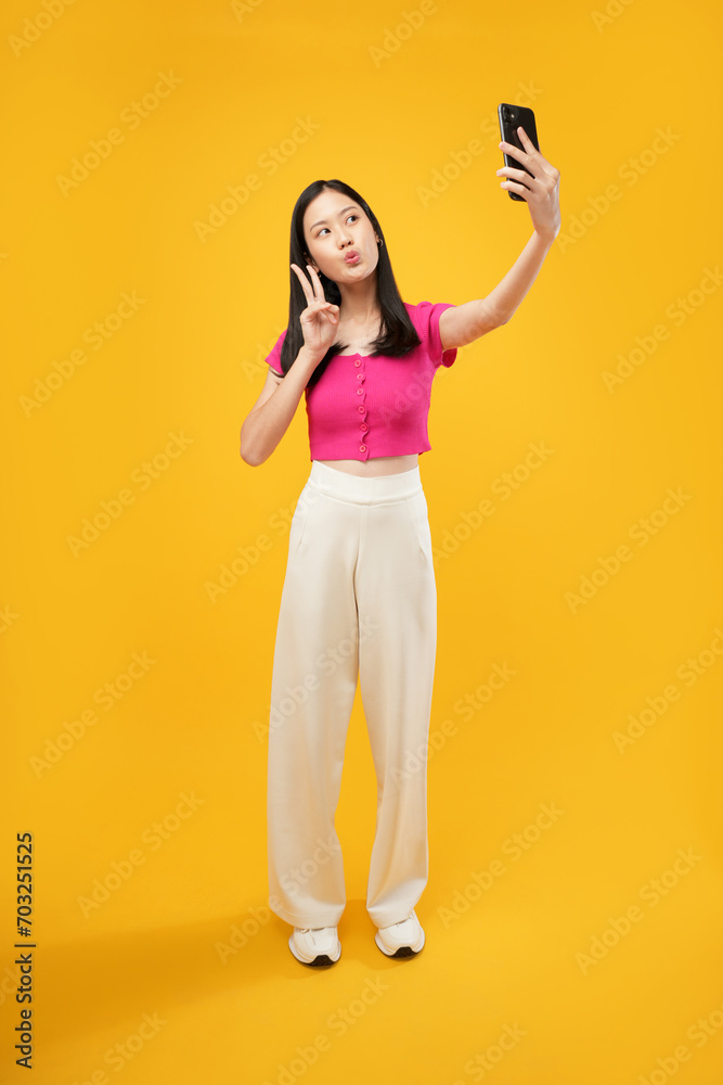 Photo of a young Asian woman holding a smartphone taking a selfie photo with a positive expression in casual clothing and looking at the camera. Isolated on a yellow background.