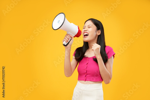 Photo of a young Asian woman holding a megaphone and shouting aggressively with fists clenched. Wearing a pink t-shirt, and isolated on a yellow background.