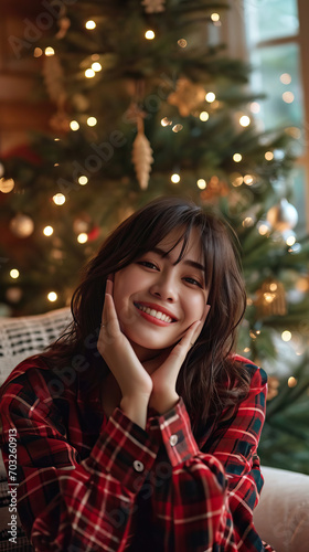 Happy Young Asian Woman Sitting on a Sofa by the Christmas Tree at Home, Embracing the Warmth and Joy of the Holiday Season