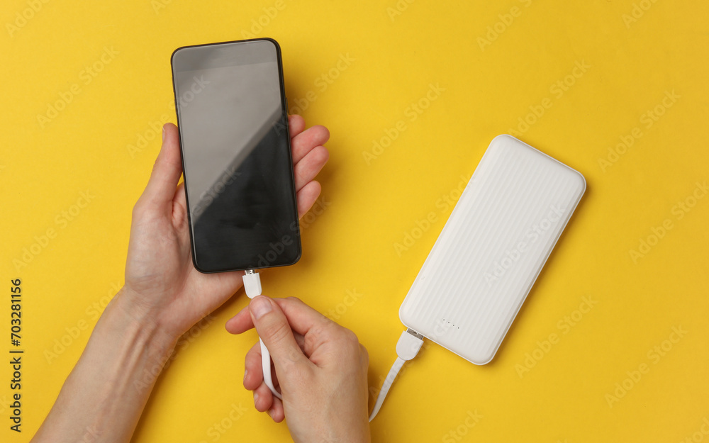 Female hands connect a smartphone to an external battery power bank on a yellow background - obrazy, fototapety, plakaty 