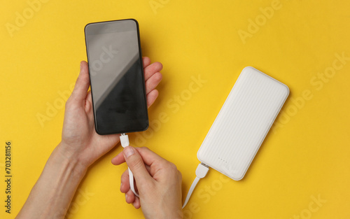 Female hands connect a smartphone to an external battery power bank on a yellow background