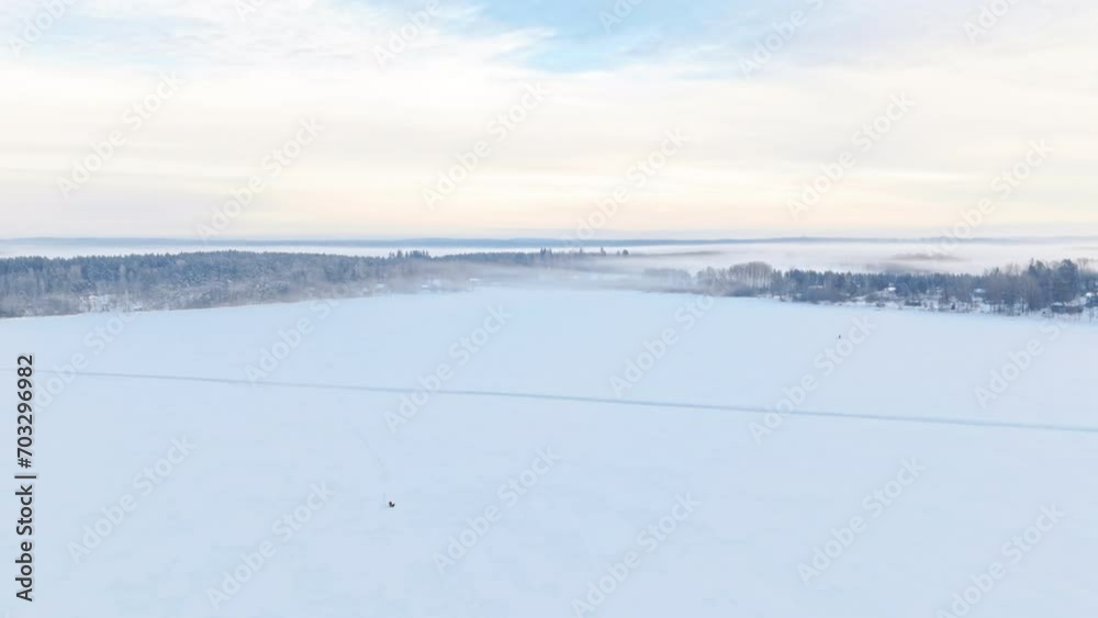 A breathtaking wide shot panorama of nordic scandinavian winter scenery, forest and lakes covered by snow and fog. A lonely ice fishing man sits on ice. Orange sky with clouds parting to reveal blue. A breathtaking wide shot panorama of nordic scandinavian winter scenery, forest and lakes covered by snow and fog. A lonely ice fishing man sits on ice. Orange sky with clouds parting to reveal blue.