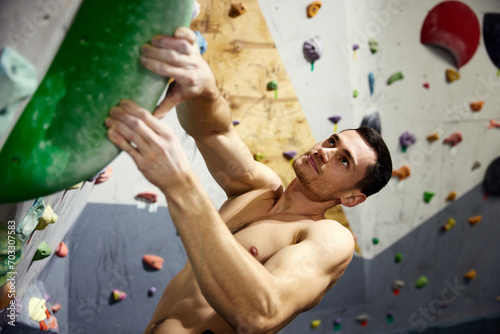 Shirtless, muscular young man climbing artificial rocks on climbing wall, practicing bouldering activity indoors. Concept of sport climbing, hobby, active lifestyle, school, training course