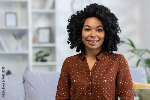 Portrait of a smiling young African American woman with curly hair sitting on the sofa at home and looking confidently at the camera. Close-up photo photo