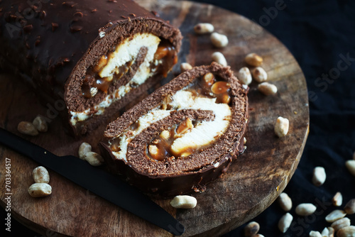 Chocolate roll with cream and caramel next to peanuts on a wooden tray on a dark background photo