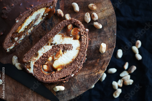 Chocolate roll with cream and caramel next to peanuts on a wooden tray on a dark background photo