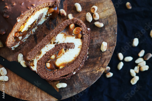 Chocolate roll with cream and caramel next to peanuts on a wooden tray on a dark background photo