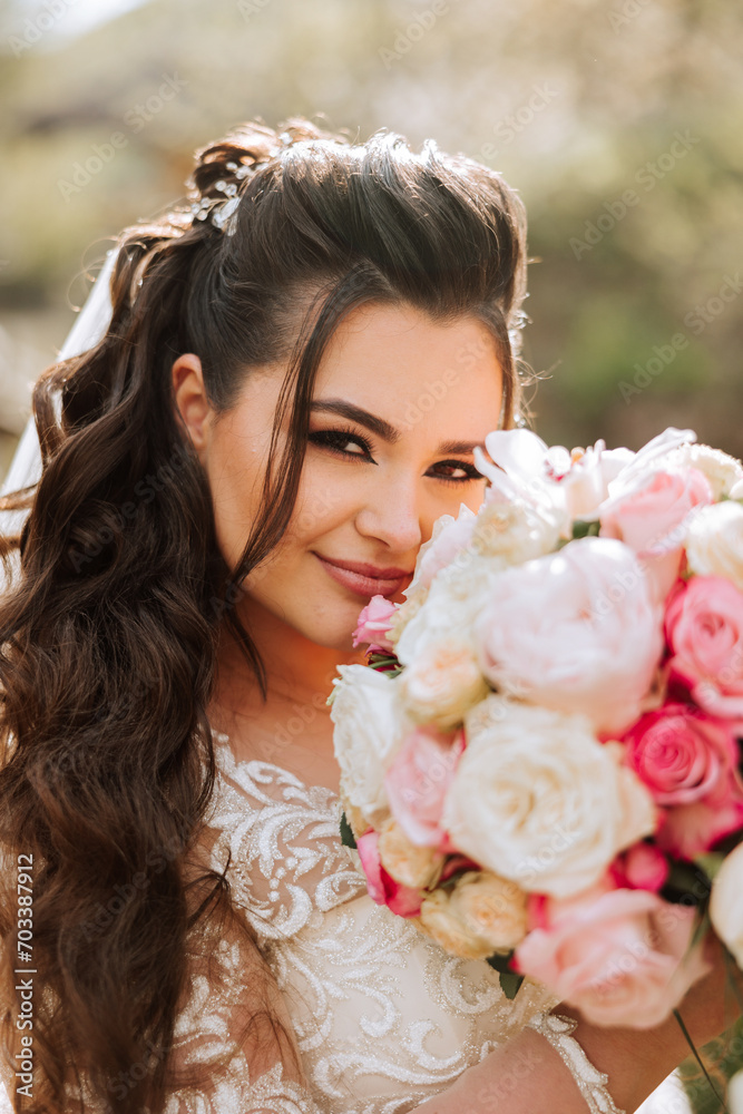 Fototapeta premium Curly brunette bride poses with a bouquet, near a wooden railing. Magnificent dress with long sleeves, open bust. Spring wedding
