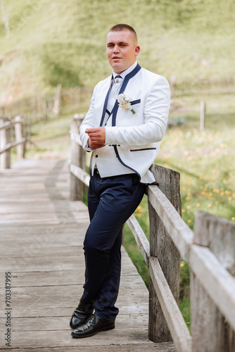The groom, in a white jacket and black pants, poses leaning on the railing and fastens a button on his sleeve. Wedding portrait.
