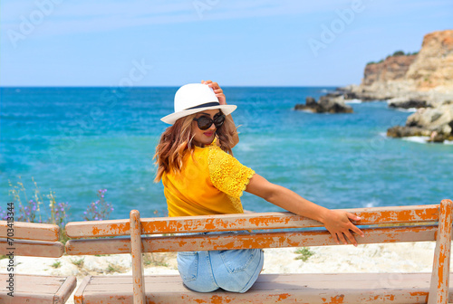 Pretty young woman in a straw hat sitting on a bench in front of the sea