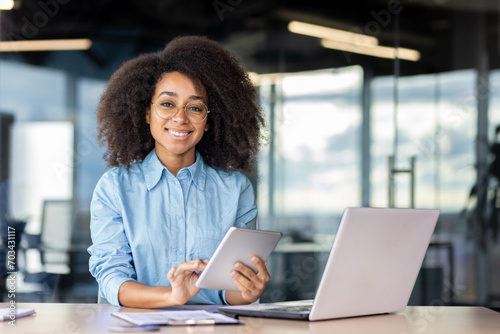 Portrait of a smiling young African American woman sitting at a desk in the office, holding a tablet and smiling at the camera. Free Download Portrait of a smiling young African American woman sitting at a desk in the office, holding a tablet and smiling at the camera. - GFXLoad