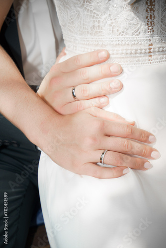 Eine männliche und eine weibliche Hand mit Eheringen auf dem weißen Brautkleid
