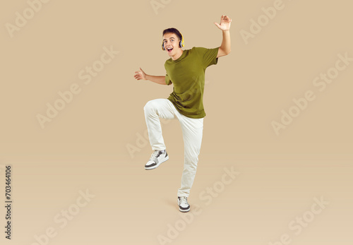 Full length portrait of a young joyful excited happy boy student wearing casual clothes having fun, dancing and listening to music in headphones isolated on a studio beige background. © Studio Romantic
